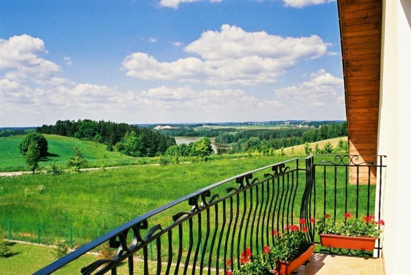 Widok z balkonu z kutą, czarną balustradą i czerwonymi pelargoniami w donicach na zielone wzgórza i lasy pod błękitnym niebem z obłokami.