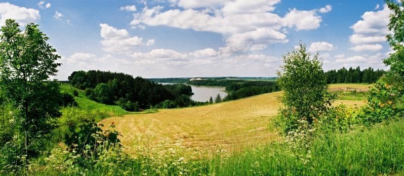 Panoramiczny widok na polodowcowy krajobraz z jeziorem w tle, widziany z zalesionego wzgórza, z polem świeżo skoszonej trawy na pierwszym planie.
