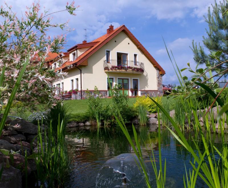 Jasny, dwupiętrowy budynek z czerwonym dachem i balkonem z kwiatami, otoczony zielenią i stawem z fontanną w słoneczny dzień.