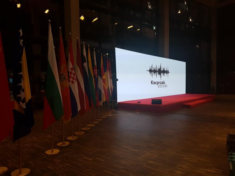 Scena z ekranem LED prezentującym logo 'Kacprzak estrada' oraz rzędem flag różnych państw, ustawiona na drewnianej podłodze.