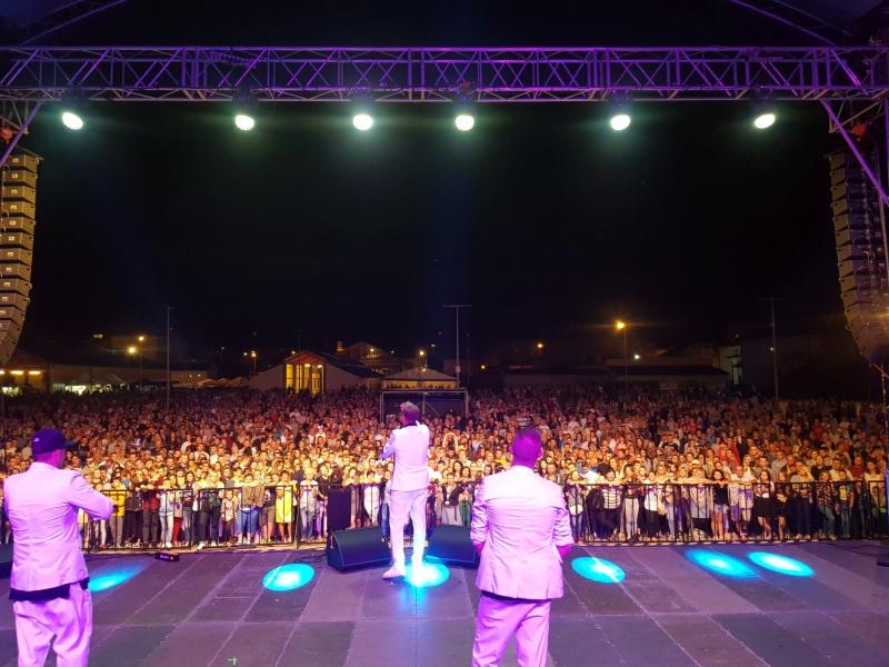 Koncert na otwartym powietrzu: artysta w białym garniturze na scenie, oświetlony fioletowym światłem, z widokiem na ogromny, rozentuzjazmowany tłum za barierkami.