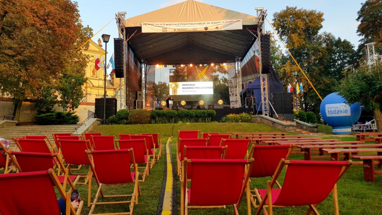Scena plenerowa z rzędami czerwonych leżaków na trawie, przygotowana na wydarzenie kulturalne, w tle budynek kościoła i baner promujący województwo łódzkie.