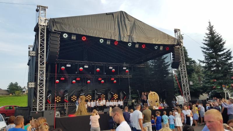 Scena plenerowa z czarnym zadaszeniem, oświetleniem i nagłośnieniem, na której występuje chór dziecięcy w białych koszulach, a po lewej stronie stoi kukła ze słomy; widoczny tłum widzów.