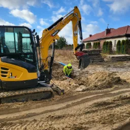 Żółta mini koparka SANY SY26U z operatorem w żółtej kurtce, który używa łopaty do kopania wąskiego wykopu w piaszczystej ziemi, na tle domu z czerwoną dachówką i niebieskiego nieba z chmurami.