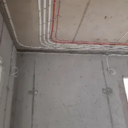 Surowe wnętrze w trakcie prac wykończeniowych: widoczne kable elektryczne prowadzone po betonie sufitu i ścian w białych listwach, częściowo z czerwonymi przewodami, z pętlą kabla zwisającą ze ściany.