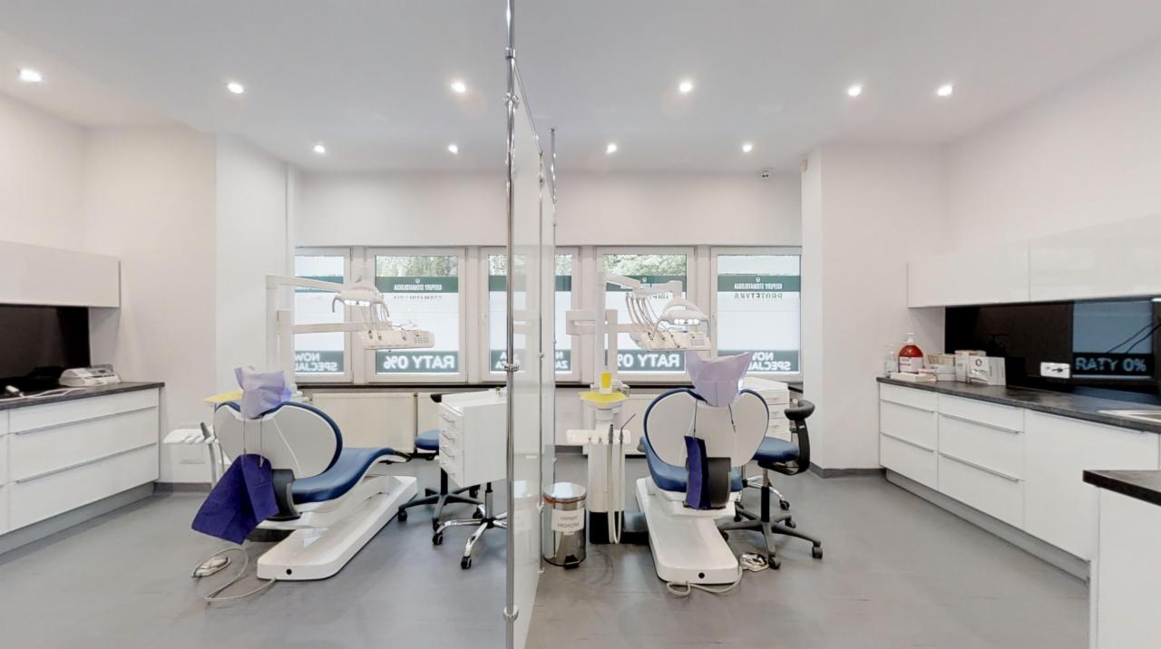 Jasne wnętrze gabinetu z dwoma stanowiskami dentystycznymi, nowoczesnym sprzętem, białymi meblami i oknami z widocznym napisem informacyjnym.