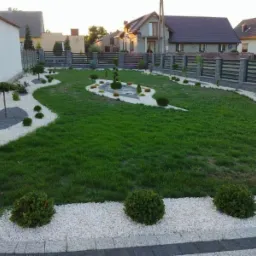 Zadbany trawnik z krzewami i drzewkami ozdobnymi, ścieżki wysypane białym grysem, nowoczesne ogrodzenie panelowe na kamiennej podmurówce w tle zabudowa jednorodzinna.