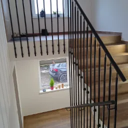 Klatka schodowa z drewnianymi stopniami i czarną, metalową konstrukcją zabezpieczającą, widok z góry na okno z małą choinką i zaparkowanym samochodem.