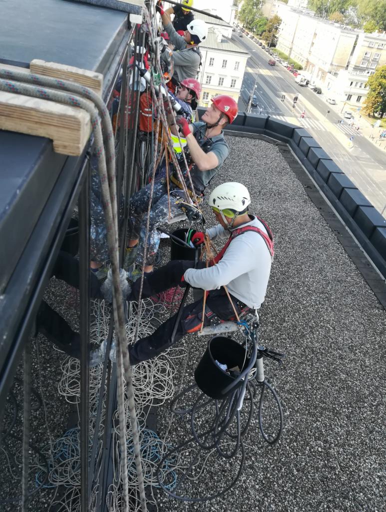 Grupa alpinistów przemysłowych w kaskach, pracujących na wysokościach przy elewacji budynku, zabezpieczonych linami, widok z góry na miasto.
