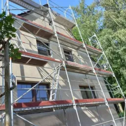 Dwupiętrowy dom w trakcie ocieplania, osłonięty rusztowaniem z metalowymi ramami i drewnianymi podestami, widoczne okna z brązowymi ramami, drzewa w tle.