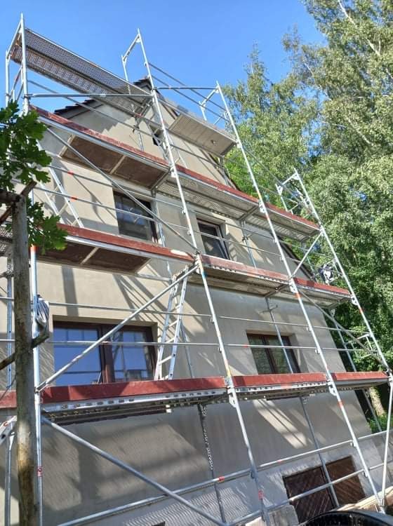 Dwupiętrowy dom w trakcie ocieplania, osłonięty rusztowaniem z metalowymi ramami i drewnianymi podestami, widoczne okna z brązowymi ramami, drzewa w tle.