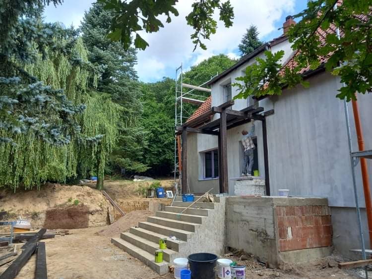 Pracownik maluje elewację domu jednorodzinnego na zewnątrz, widoczne rusztowanie, przygotowany teren i częściowo pomalowana ściana. Nowe schody prowadzą do wejścia.