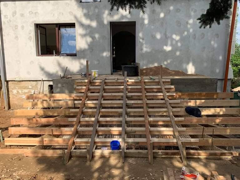 Budowa betonowych schodów wejściowych do domu, widoczna drewniana konstrukcja szalunkowa i zbrojenie, ściana budynku w trakcie tynkowania.