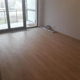 Ułożone panele podłogowe w pustym pokoju z balkonem, widoczne listwy przypodłogowe i wiaderko farby przy ścianie.