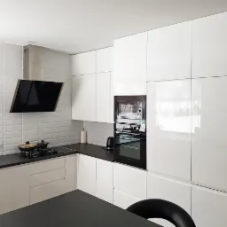 Minimalistyczna kuchnia z białymi, lakierowanymi frontami, czarnym blatem i czarnym, skośnym okapem nad płytą grzewczą z dwoma garnkami.