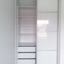 Biała szafa wnękowa z przesuwnymi drzwiami z mlecznego szkła, widoczne półki i szuflady, drewniana podłoga.