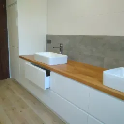 Nowoczesna łazienka z drewnianym blatem, dwiema prostokątnymi umywalkami nablatowymi i białymi szafkami z wysuwaną szufladą, uzupełniona płytkami w odcieniach szarości i bieli.