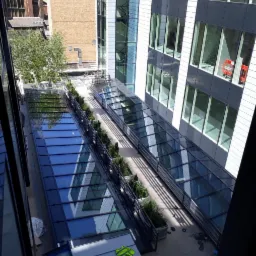 Wewnętrzny dziedziniec biurowca z przeszklonymi zadaszeniami, widok z góry, zieleń w donicach i refleksy w szkle.