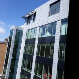 Nowoczesny budynek biurowy z fasadą z paneli w kolorze jasnoszarym i dużymi oknami z czarnymi ramami, widok z dołu w słoneczny dzień.