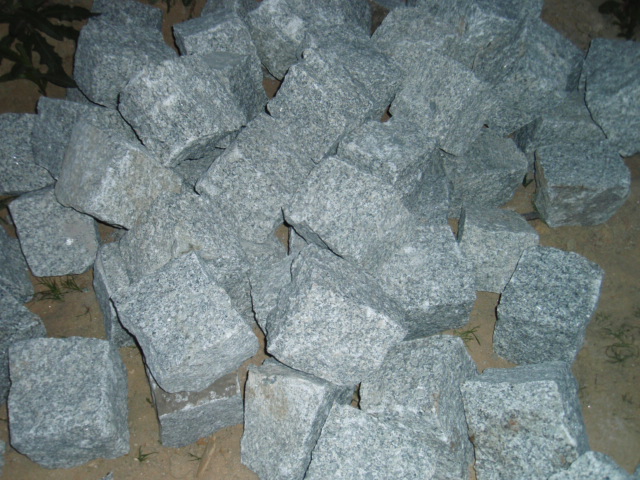 Stos szarych, surowych kostek z granitu o nieregularnych kształtach, ułożonych na ziemi, z widocznymi drobnymi źdźbłami trawy.