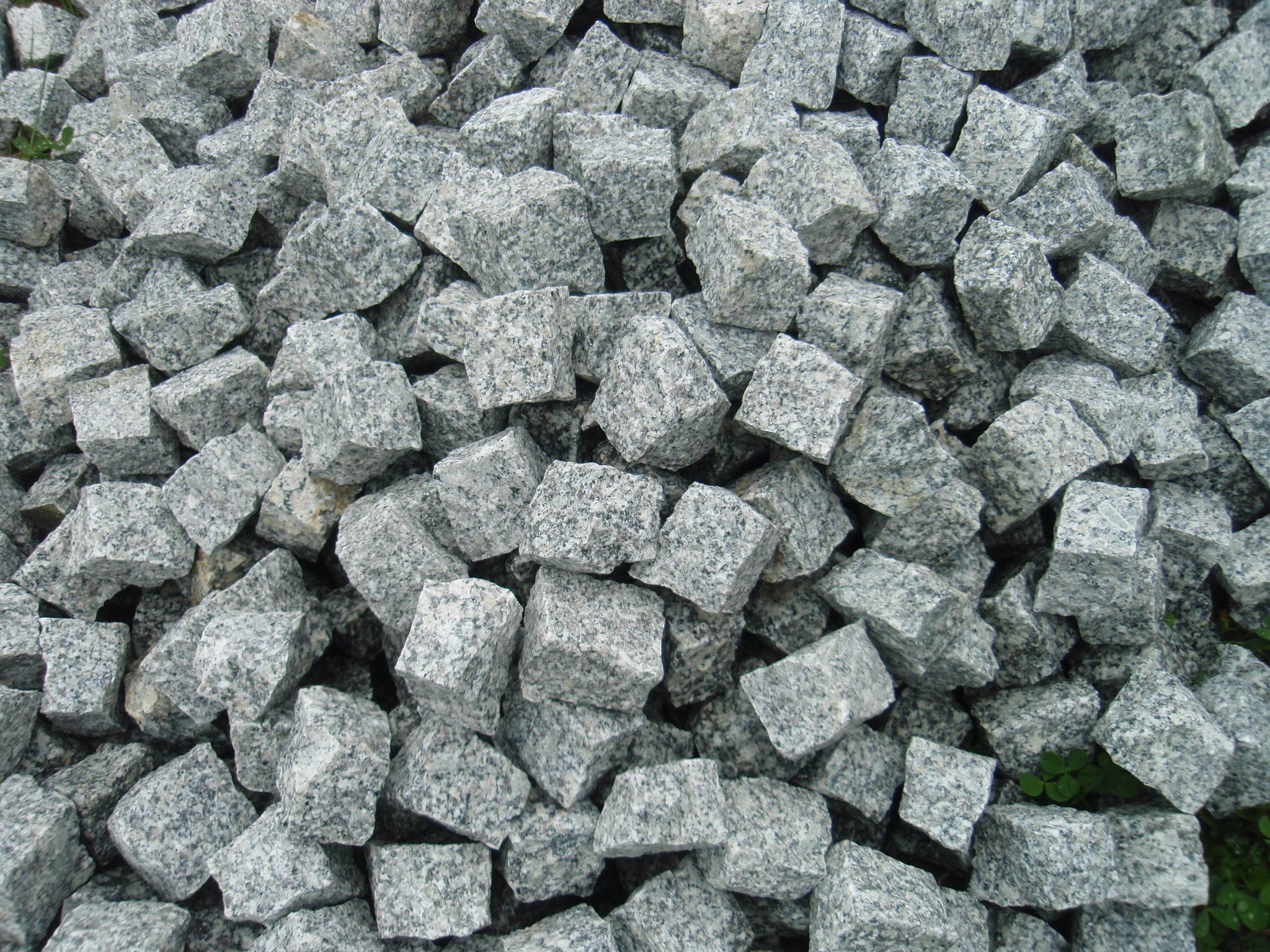 Kupa szarej, gruboziarnistej kostki o nieregularnych kształtach, z niewielkimi fragmentami zielonej trawy widocznymi w tle.