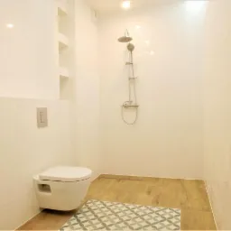 Nowoczesna łazienka z białą armaturą, prysznicem bez brodzika i drewnopodobną podłogą, akcentowana wzorzystym dywanikiem.