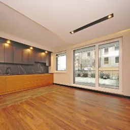 Nowoczesna kuchnia z drewnianymi szafkami dolnymi i ciemnoszarymi górnymi, marmurowy backsplash, parkiet i duże okno przesuwne z widokiem na ogród w tle.