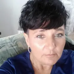 Portret kobiety z krótkimi, ciemnymi włosami i srebrnymi kolczykami, ubrana w niebieską koszulkę. W tle widoczny fragment ściany z cegły i dekoracyjny element.