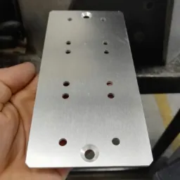 Aluminiowa płytka z nawierconymi otworami, trzymana w dłoni, widoczne ślady obróbki skrawaniem.