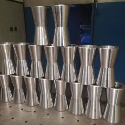 Stos obrobionych tokarsko, aluminiowych elementów o kształcie klepsydry, ułożonych piramidalnie na metalowym stole spawalniczym z widocznymi otworami i napisem, w tle ściana z ostrzeżeniem.