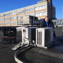 Instalator w kurtce i niebieskiej koszuli sprawdza klimatyzator Panasonic na dachu, obok skrzynka z narzędziami i kable w peszlach.