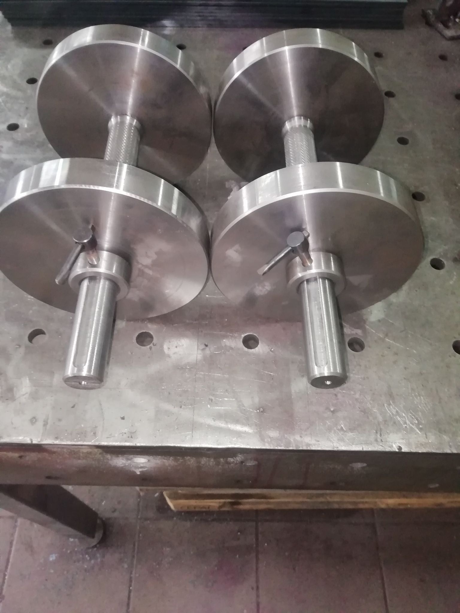 Dwie pary metalowych hantli z okrągłymi obciążeniami leżące na metalowym stole warsztatowym z otworami.