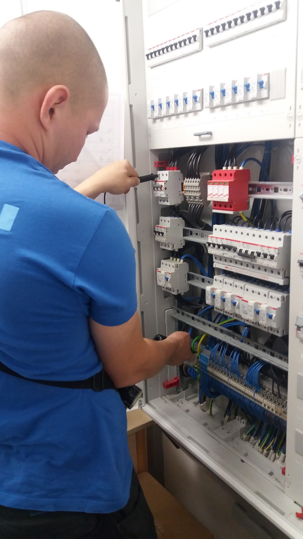 Elektryk w niebieskiej koszulce sprawdza napięcie w rozdzielnicy elektrycznej za pomocą miernika, widoczne liczne kable i zabezpieczenia.