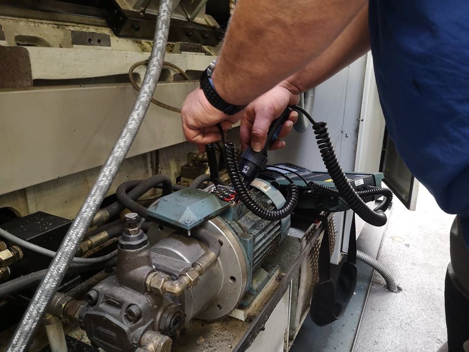 Mężczyzna w niebieskiej koszulce dokonuje pomiarów na silniku maszyny, używając specjalistycznego urządzenia z kablem spiralnym.