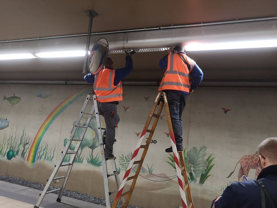 Dwóch mężczyzn w kamizelkach odblaskowych na drabinach, montujących oświetlenie sufitowe w tunelu z malowidłami ściennymi.