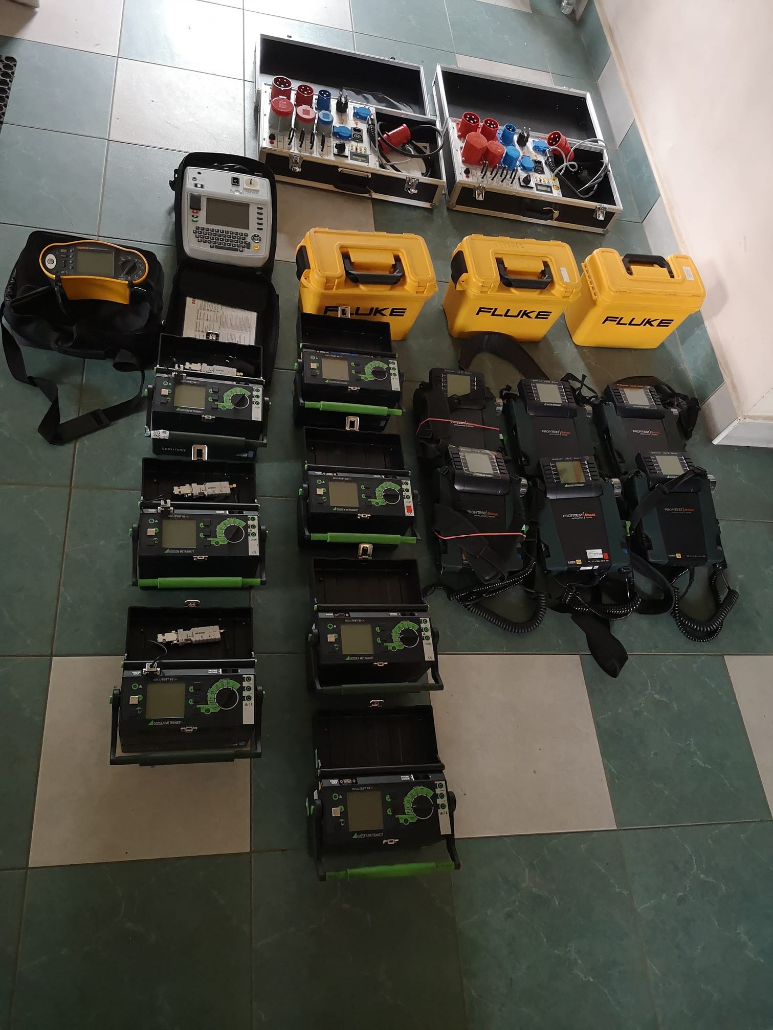 Zestaw narzędzi i urządzeń pomiarowych, w tym testery instalacji elektrycznych marek Fluke i Gossen Metrawatt, rozłożony na podłodze z płytek, gotowy do użycia w terenie.
