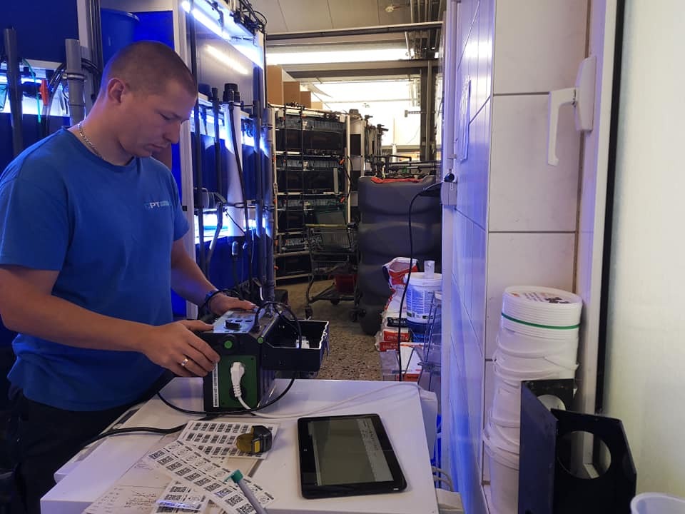 Mężczyzna w niebieskiej koszulce z logo 'PT' konfiguruje urządzenie elektroniczne z zielonym gniazdem, na stole tablet i etykiety, w tle regały z oświetlonymi zbiornikami.