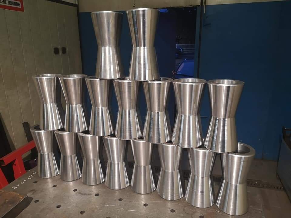 Stos obrobionych tokarsko, aluminiowych elementów o kształcie klepsydry, ułożonych piramidalnie na metalowym stole spawalniczym z widocznymi otworami i napisem, w tle ściana z ostrzeżeniem.