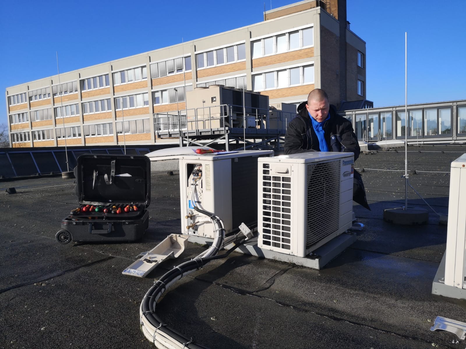 Instalator w kurtce i niebieskiej koszuli sprawdza klimatyzator Panasonic na dachu, obok skrzynka z narzędziami i kable w peszlach.