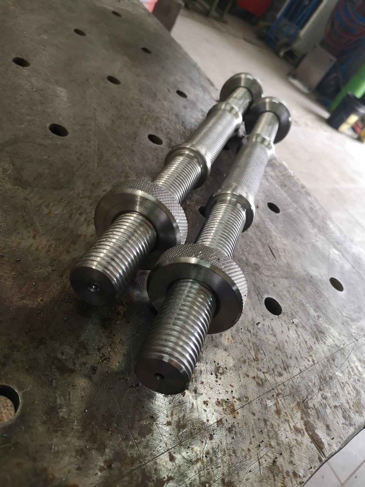 Dwa metalowe pręty z gwintem i radełkowaną powierzchnią na metalowym stole z otworami, widoczne ślady obróbki.