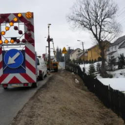 Prace wiertnicze przy drodze z użyciem maszyny wiertniczej i pojazdu z sygnalizacją świetlną, w tle domy mieszkalne i resztki śniegu.