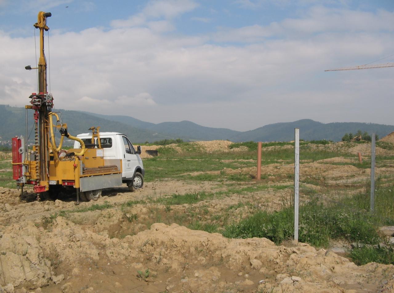 Wiertnica zamontowana na samochodzie dostawczym, prowadząca prace wiertnicze na terenie budowy z widokiem na góry w tle.