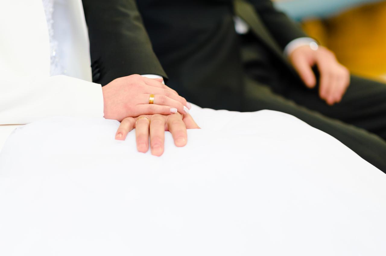 Dłonie nowożeńców spoczywają na białej sukni, widoczne złote obrączki na palcach.