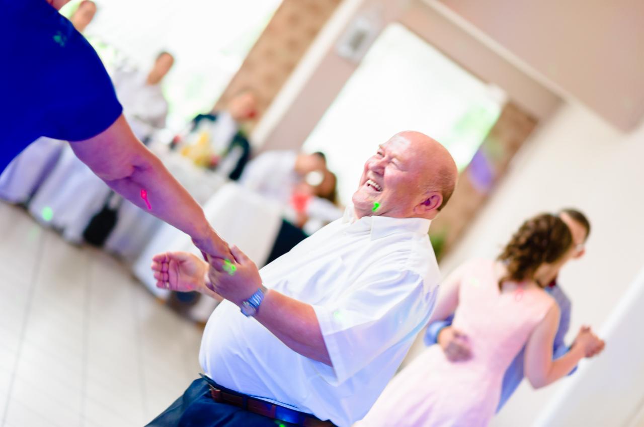 Rozbawiony mężczyzna w białej koszuli trzymający za ręce inną osobę podczas tańca na weselu, w tle para tańcząca w objęciach.