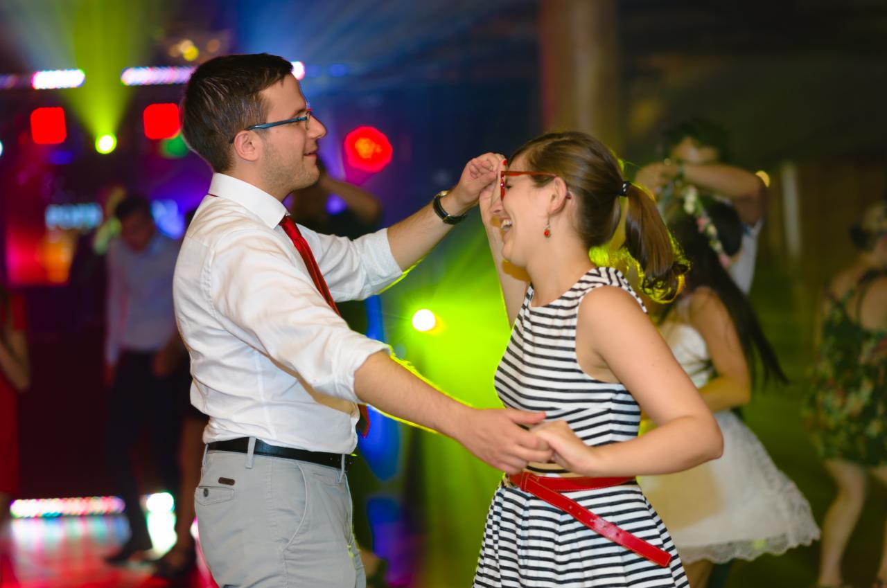 Para młoda tańczy na parkiecie podczas wesela, mężczyzna w okularach poprawia kobiecie okulary, w tle kolorowe światła i inni goście.