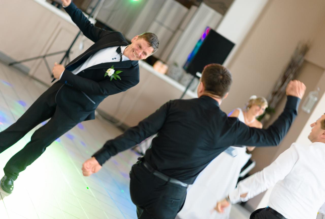 Rozbawiony pan młody w czarnym garniturze tańczy z gośćmi na parkiecie oświetlonym kolorowymi światłami podczas wesela, w tle uśmiechnięta panna młoda.