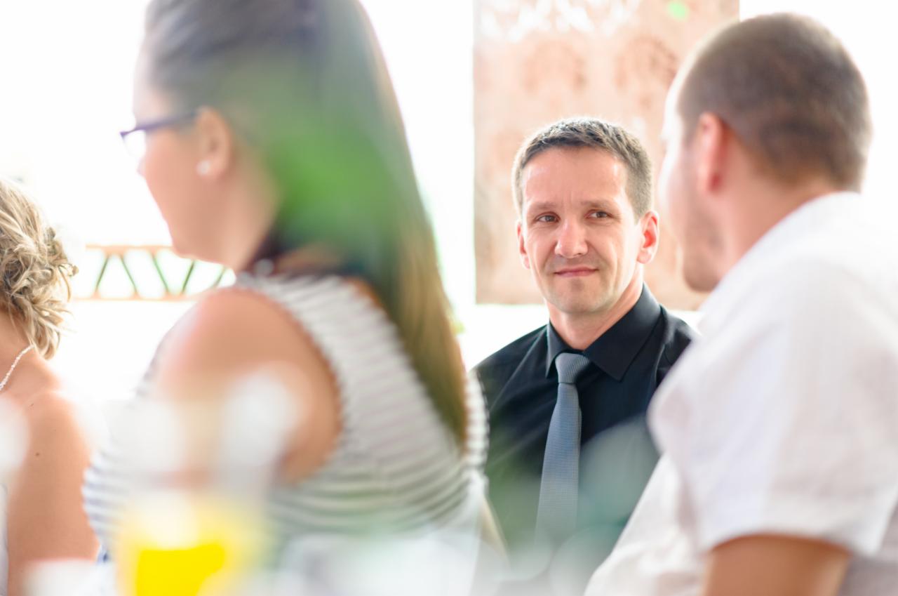 Mężczyzna w czarnej koszuli i krawacie spogląda z uśmiechem przez ramię, otoczony częściowo rozmytymi postaciami podczas wydarzenia.