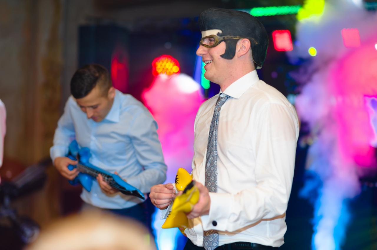 Mężczyzna w przebraniu Elvisa z plastikową gitarą na imprezie, drugi mężczyzna w tle gra na niebieskiej, dmuchanej gitarze, kolorowe światła i dym.