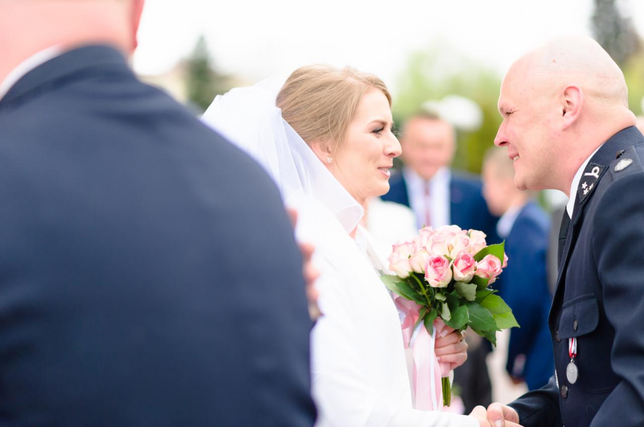 Panna młoda w białej sukni i welonie trzyma bukiet różowych róż, uśmiecha się do mężczyzny w mundurze, ceremonia ślubna z udziałem wojskowego.