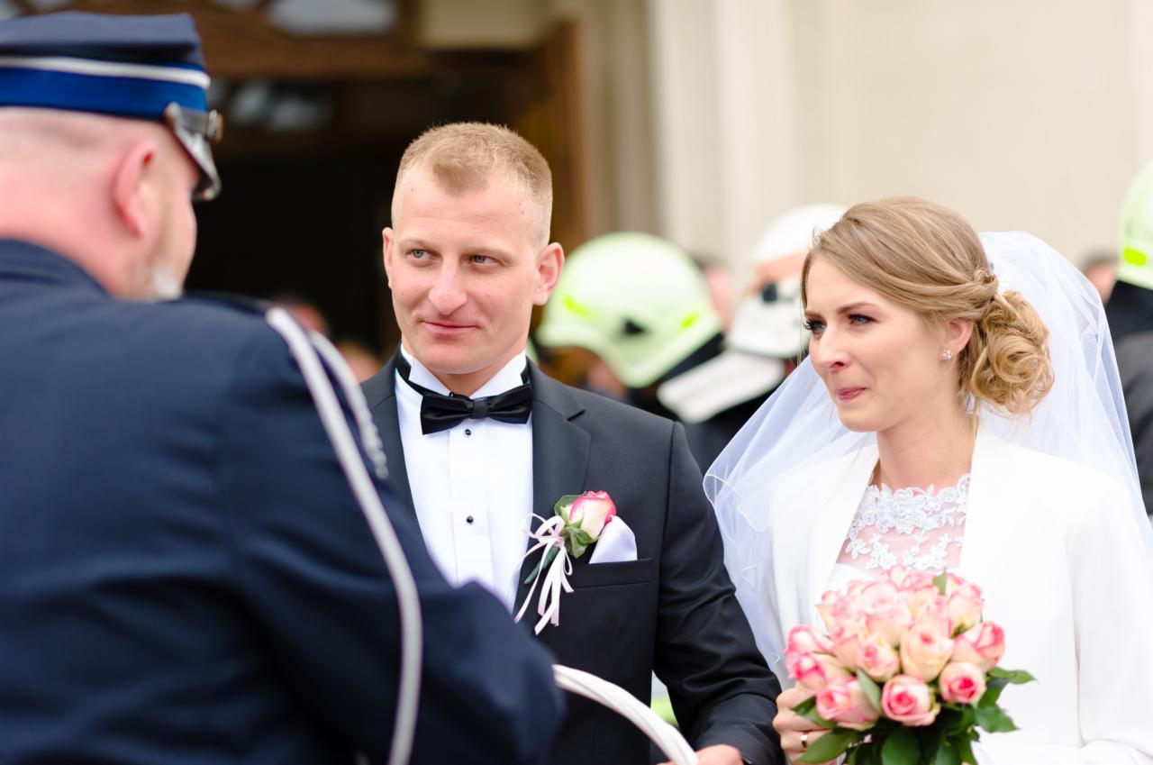 Uśmiechnięty pan młody w czarnym smokingu z różą w butonierce, panna młoda w białej sukni i welonie trzyma bukiet różowych róż, w tle strażacy w hełmach.